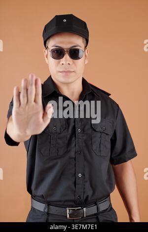 Serious Asian male security officer making stop gesture standing against orange background in studio Stock Photo