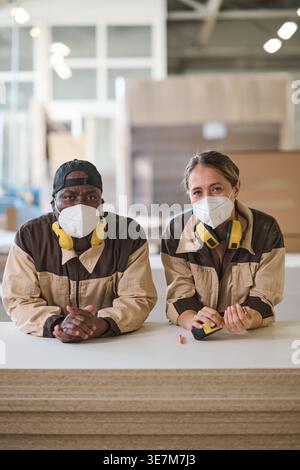 Middle-aged Black male manual worker and caucasian female coworker wearing face masks leaning on wooden planks in warehouse Stock Photo
