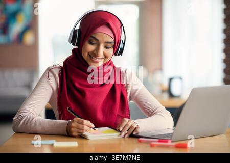 Indian Muslim woman in hijab wearing headphones taking notes sitting at table with laptop in cafe Stock Photo