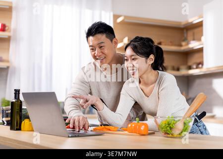 Asian couple using laptop together while bending forward at table with vegetables at home Stock Photo