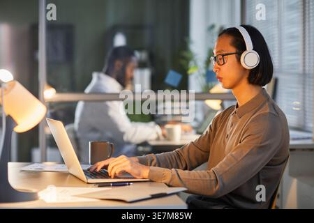 Asian man wearing headphones using laptop at workplace working late in office lit by lamps Stock Photo