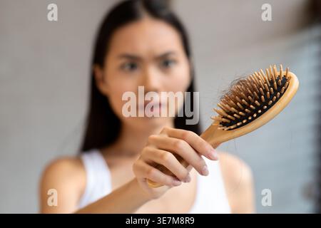 Worried long-haired millennial asian woman holding hairbrush, closeup. Young korean lady loosing hair, checking brush after combing hair. Hair loss, a Stock Photo