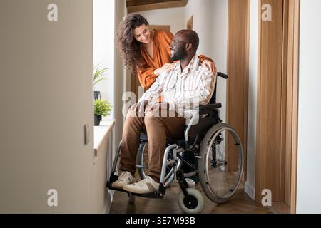 Black bald man sitting in wheelchair talking to caucasian girlfriend in corridor at home Stock Photo