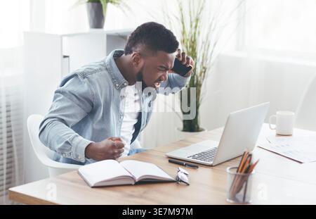 Angry Black businessman having phone call looking  at laptop sitting with closed fist at table with notebook in office Stock Photo