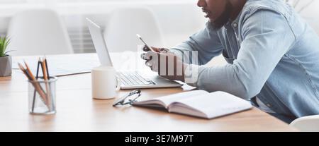 Black businessman using smartphone sitting at table with laptop in office Stock Photo