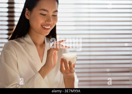 Asian woman wearing bathrobe holding jar with moisturizing cream while standing in bathroom at home Stock Photo