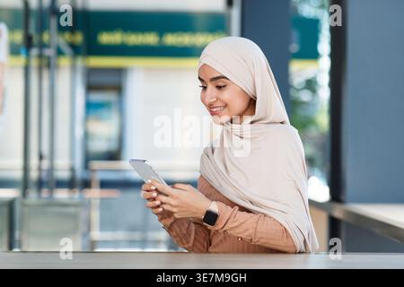 Smiling Indian Muslim woman wearing hijab using smartphone sitting at table in outdoor cafe during daytime Stock Photo