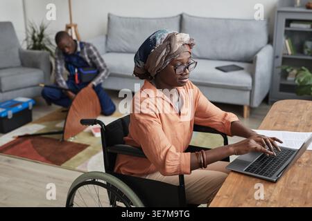 Disabled Black woman wearing head wrap and eyeglasses working on laptop while sitting in wheelchair near handyman assembling furniture in living ro... Stock Photo