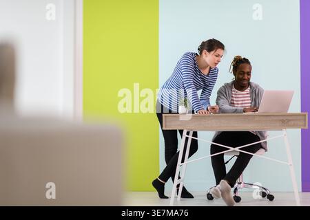 Smiling Black businessman with braids sitting at table working on laptop and having discussion with caucasian female colleague standing beside him ... Stock Photo