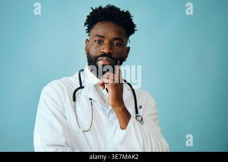 Bearded Black male doctor with stethoscope around his neck standing with hand on face against turquoise background in studio Stock Photo