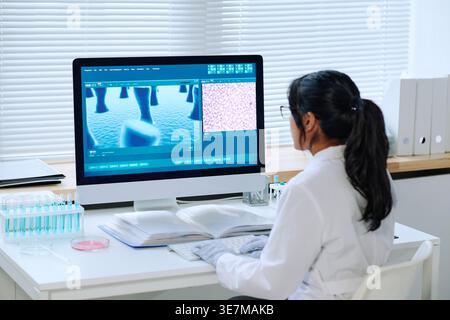 Hispanic woman wearing white lab coat and eyeglasses studying image on computer screen sitting at table in laboratory Stock Photo