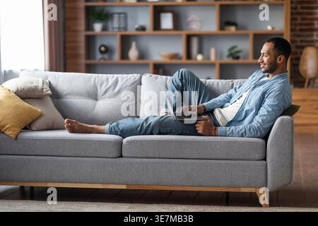 Black man using laptop while lying on sofa in living room at home Stock Photo