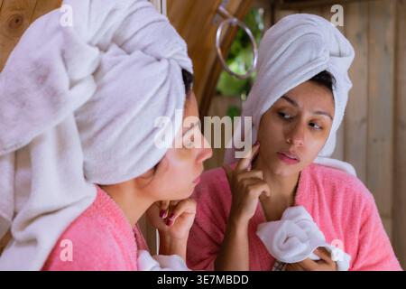 Hispanic woman with head wrapped in towel pointing finger on face standing in front of the mirror at home Stock Photo