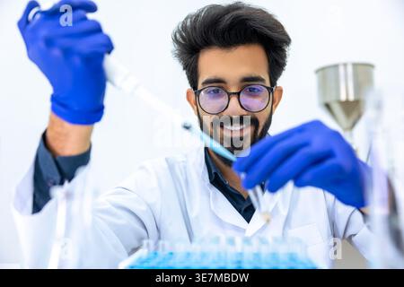Smiling Indian male scientist wearing lab coat and latex gloves dripping chemicals to test tubes conducting experiments in science lab Stock Photo