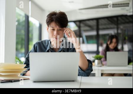 Stressed Asian male office worker working on laptop sitting at desk in office Stock Photo