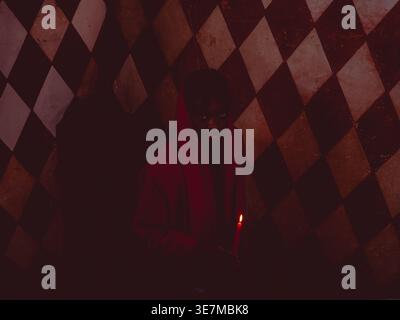 Black woman with short hair wearing red suit holding lit candle standing on stairs with argyle wall in dark house during halloween Stock Photo