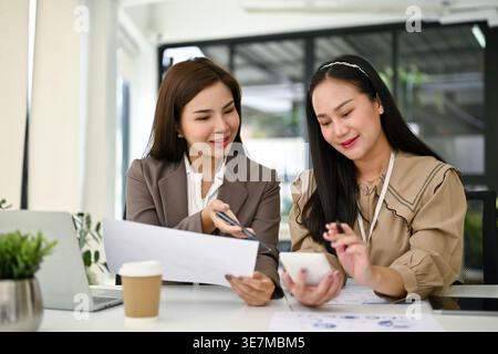 Smiling Asian female accountants working and analyzing financial data reports while sitting together at table in office Stock Photo