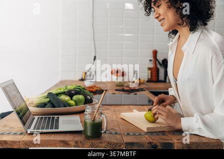 Hispanic woman cutting lime with knife looking at laptop standing in kitchen Stock Photo