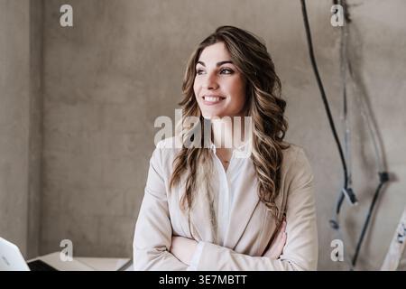 Smiling Hispanic female architect with crossed arms looking away standing against table in construction site during daytime Stock Photo