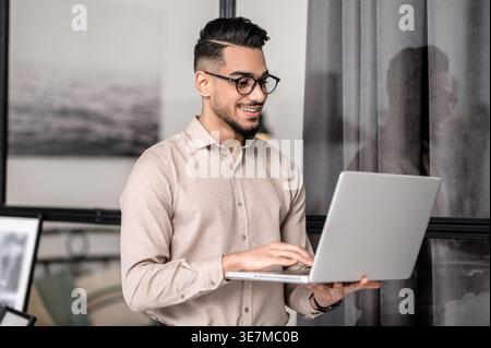 Smiling Arab man wearing eyeglasses standing while using laptop at home Stock Photo