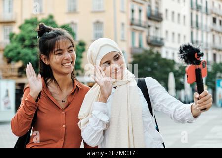 Middle Eastern and Asian female friends waving hands streaming using smartphone standing on city street during daytime Stock Photo