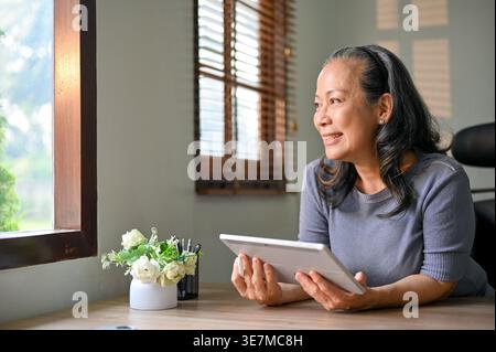 Smiling senior Asian woman holding tablet while leaning and sitting at table in home office Stock Photo
