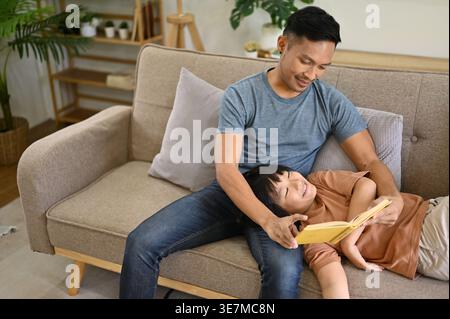 Smiling Asian boy lying on lap of his father on sofa and reading book at home Stock Photo