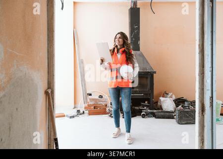 Smiling Hispanic female architect holding clipboard and hard hat looking away standing in construction site during daytime Stock Photo