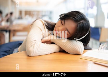 Sleeping Asian woman listening to music with headphones leaning on table sitting in cafe Stock Photo