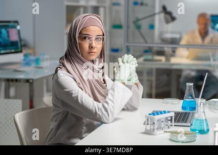 Serious Mixed race female scientist in hijab wearing protective glasses with clasped hands in the laboratory clinic Stock Photo