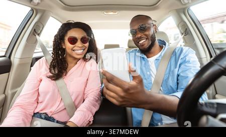 Smiling Black couple with sunglasses sharing smartphone while riding car together in city Stock Photo