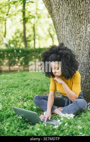Hispanic female with curly hair leaning chin on hand working on laptop sitting against tree trunk in park during daytime Stock Photo