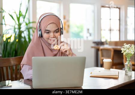 Asian Muslim woman wearing hijab using laptop with headphones while eating cookies sitting at table in cafe Stock Photo