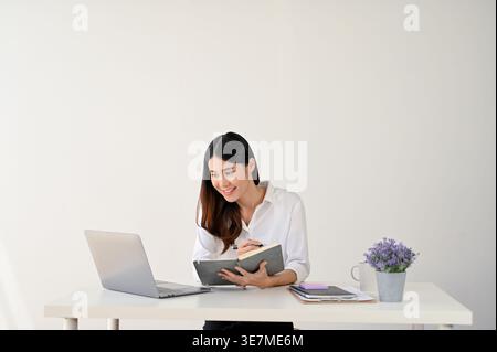 Smiling Asian business woman taking down notes while looking at laptop and sitting at table against white background Stock Photo