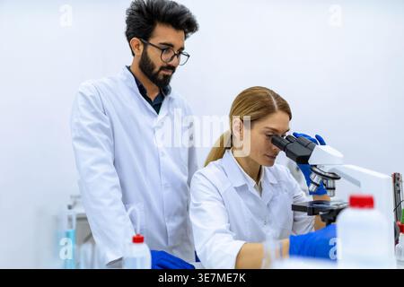 Indian male and caucasian female scientists wearing lab coats and latex gloves using microscope doing sample analysis together in laboratory Stock Photo