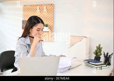 Asian business woman reading and checking financial reports while sitting at table with laptop in office Stock Photo