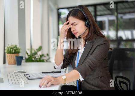Stressed Asian female support agent with headset having headache touching forehead sitting at table while working in office Stock Photo