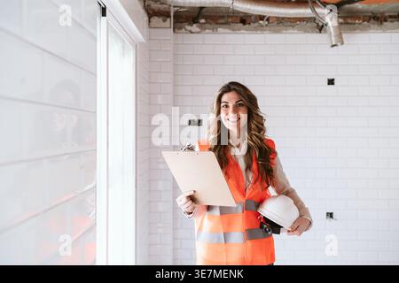 Smiling Hispanic female architect wearing safety vest holding clipboard and hard hat standing in construction site during daytime Stock Photo