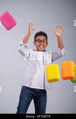 Cheerful African American boy with raised arms standing near flying food containers against wall Stock Photo