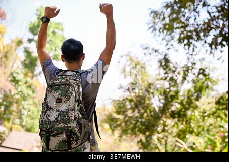 Asian male traveler raising arms while standing with backpack in forest on sunny day Stock Photo