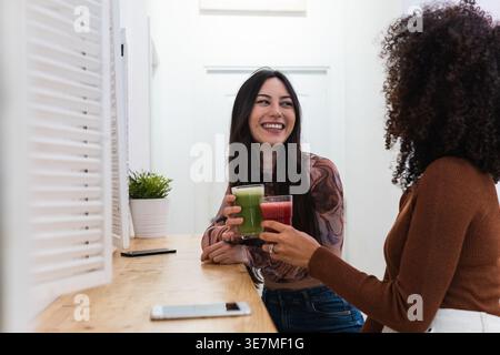 Mixed race and Hispanic female friends looking at each other and clinking glasses of smoothies while sitting together at wooden table in cafe Stock Photo