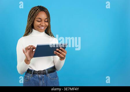 Smiling Black female with braids using tablet while standing against blue background Stock Photo