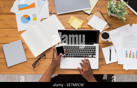 Black businessman using smartphone and laptop sitting at wooden desk various objects in office Stock Photo