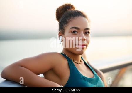 Smiling Mixed race woman in wireless earphones leaning arms on railing while standing on promenade during daytime Stock Photo
