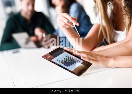 Middle Eastern and caucasian women working together on tablet near colleagues having a meeting while sitting at table in office Stock Photo