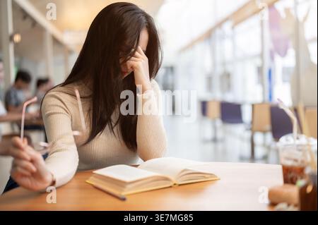 Tired Asian woman  suffering from eye strain takes off her glasses after long reading session sitting at table in cafe Stock Photo