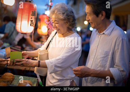 Smiling senior Chinese couple buying street foods while standing together in street market at night Stock Photo