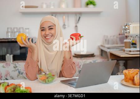 Smiling Asian Muslim woman in hijab sitting with both arms on table near laptop while holding peppers in kitchen at home Stock Photo