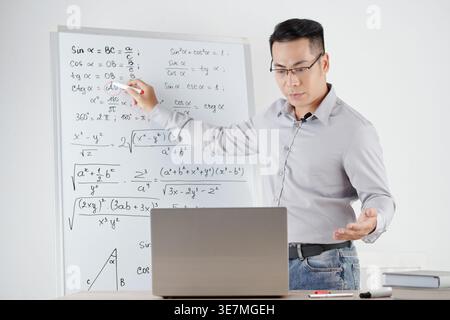 Serious Asian male mathematics teacher answering question of online students and explaining every variable in formula in classroom Stock Photo