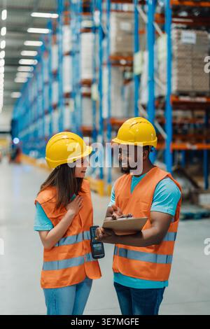 Black male worker wearing hard hat and safety vest holding clipboard and managing inventory with caucasian female coworker while standing in warehouse Stock Photo